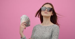 Arrogant young woman in trendy sunglasses holding fan made of dollars banknotes while posing in studio. Beautiful girl with dark hair enjoying rich life over pink background.