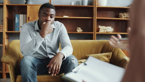 Selective focus shot of unhappy African American man sitting on couch in counseling office and complaining about his troubles to female psychologist while she taking notes on clipboard