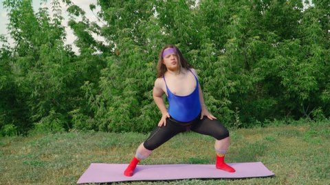 Funny Fat Man with Long Hair in a Tight Female T-shirt Is Engaged in Martial Arts on fitness mat on Green Lawn in City Park. Playful Guy Overweight Depicting a Girl. Sport humor concept.