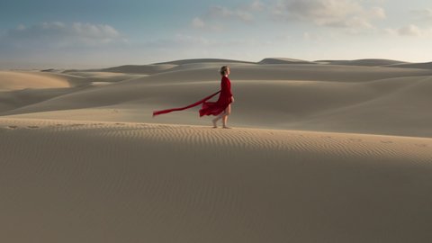 Nature travel aerial video. Young female in a beautiful wavy red dress walking through the natural landscape. 4K Aerial drone footage of a caucasian woman at a windy day, on the sandhill at sunset