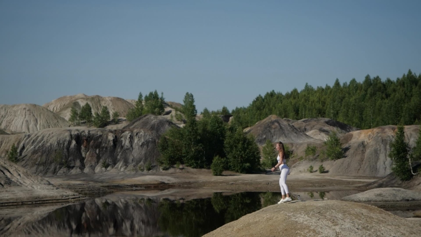 A girl makes lunges with a jump up the lake in a career. The athlete in white uniform trains against the background of sandy mountains and forests in the Urals.