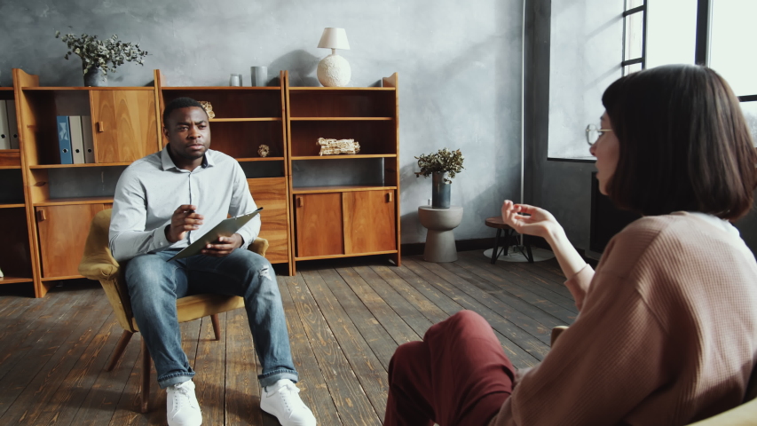 Over the shoulder shot of African American male psychologist sitting in armchair, taking notes on clipboard and listening attentively to female patient during counseling session in his office