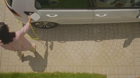 High angle shot of a woman plugging the eco-friendly electric car to charge on her backyard