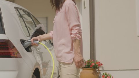A woman walking up to her electric vehicle, plugging it in to charge in front of her house