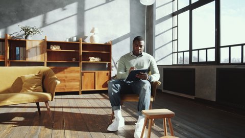 Zoom in shot of young African American male counselor sitting in armchair, writing on clipboard and looking through window thoughtfully while working in his office