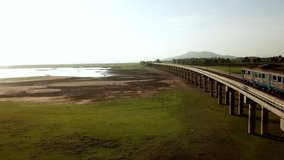 Aerial view of passenger diesel train running on a railway bridge above grassland across the lake of Pa Sak Jolasid dam in Lopburi, Thailand during morning sunrise. This clip is have noise or grain.