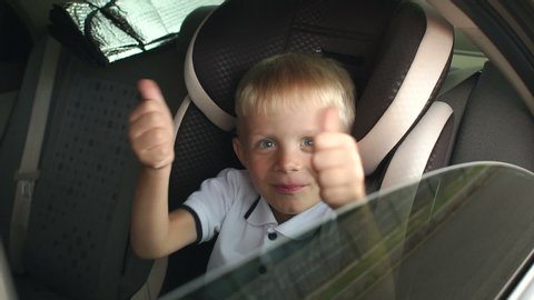 A small cheerful boy sits in a brown child car seat and shows his thumb up. Cute kid is playing and having fun in the kids car seat during the summer journey, slow motion. Child transportation safety.