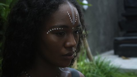Video of a beautiful young brunette African girl with makeup and painted ethnic lines on the body sitting in a tropical garden