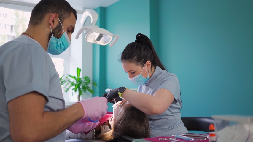 Two doctors, man and woman in protective masks in a dental clinic, serve the patient girl. Work Dentist. Healthcare and medicine concept. Steadicam shooting. | Shutterstock HD Video #1055304500