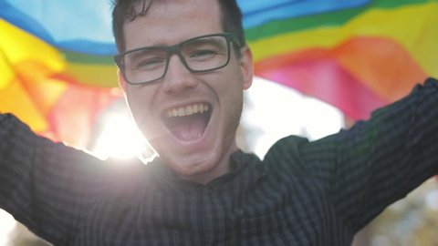 Portrait of cheerful millennial guy with earings looking to camera. Close up view of young handsome man with long wavy hair smiling, posing with rainbow flag at background. Concept of pride