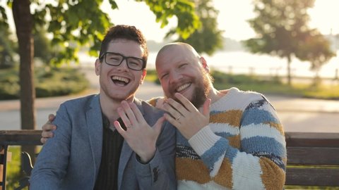 Gay Marriage Proposal Concept. Adorable Boyfriend show his Beautiful Shiny Wedding Rings to camera. Surprised Partner is Extremelly Happy and Hugs His Queer Friend. Relationship Goals.