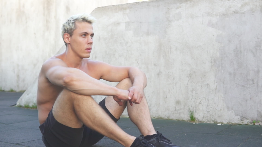 Young caucasian tired athlete man wipes sweat from forehead sitting on work out area outdoors