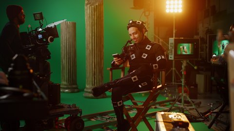 Handsome Smiling Actor Wearing Motion Capture Suit and Head Rig having Lunch Break, Sitting on Chair, Uses Smartphone. Studio High Budget Movie. On Film Studio Period Costume Drama Film Set