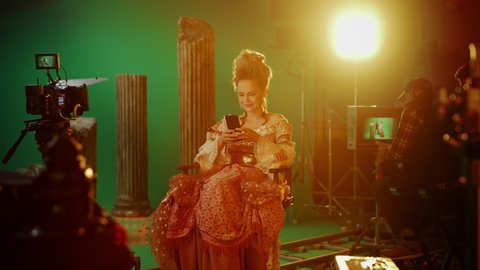 On Period Costume Drama Film Set: Beautiful Smiling Actress Wearing Renaissance Dress, Sitting on a Chair Using Smartphone with Green Screen in the Background. Studio Shooting Historical Blockbuster