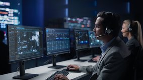 Confident Male Cyber Security Specialist Wearing Headset Works on Personal Computer Showing Footage of Drone Surveillance Tracking of a Vehicle. Big Monitoring Room with Professional People Working - Powered by Shutterstock - Get 15% off with code: PIKWIZARD15