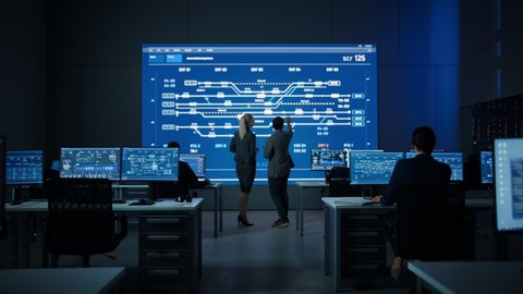 Project Manager and Computer Science Engineer Talking while Using Big Screen Display Showing Infrastructure Infographics, Data. Telecommunications Company System Control and Monitoring Room. Zoom out