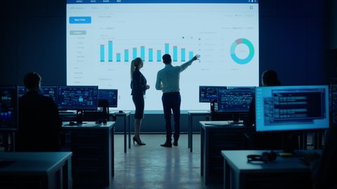 Project Leader and Chief Executive Discuss Data Shown on Big Display. Screens Show Infographics, Charts, Finance Analysis, Stock Market, Growth.Telecommunications Control Room Working Professionals