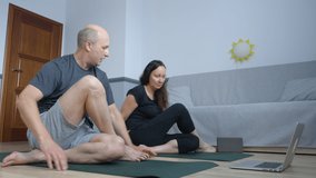 Adult sport couple practicing twist exercise yoga asana front laptop in home. Flexible man and woman training together yoga exercise online on floor in home.