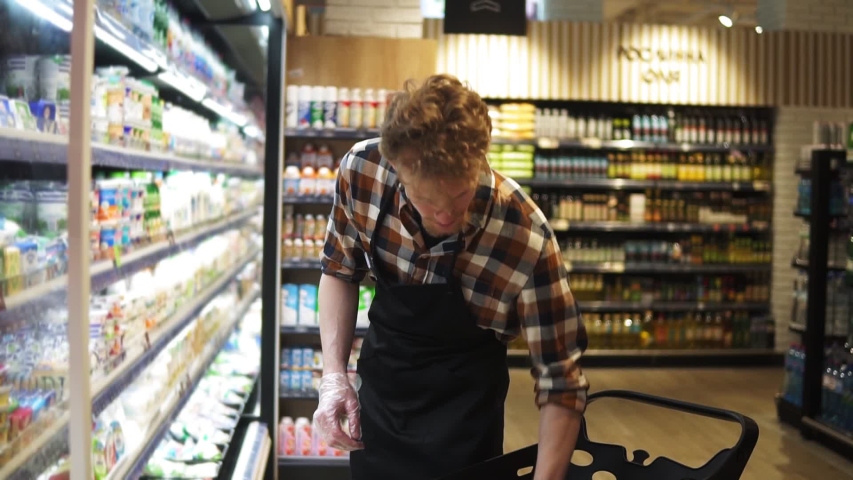 Salesman is putting cheese packs from the cart on shelves in milk products department in food store, bearded guy is wearing apron. Selling products, profession and trade concept Royalty-Free Stock Footage #1055323700