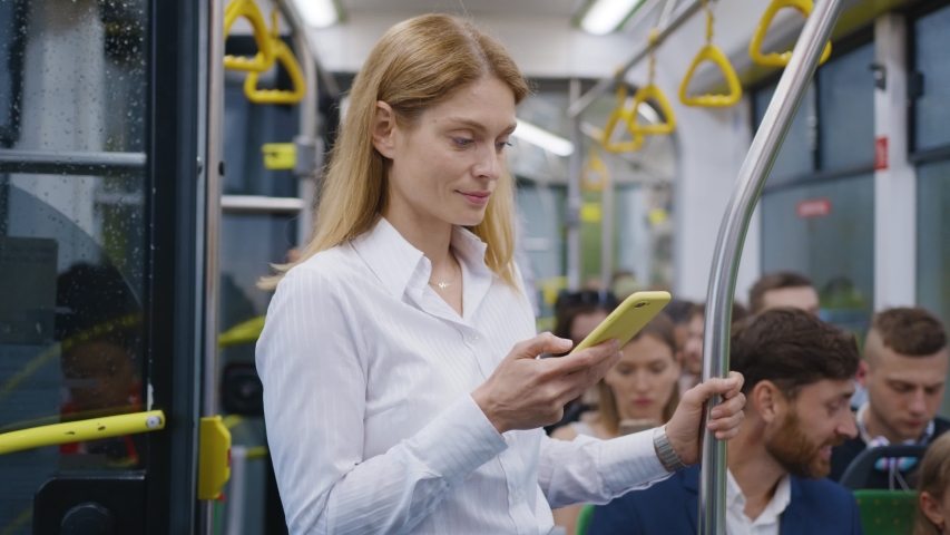Nice-looking blonde business woman browsing social media news, smiling while reading on smartphone screen, standing in the bus. Public transport. Internet addiction.