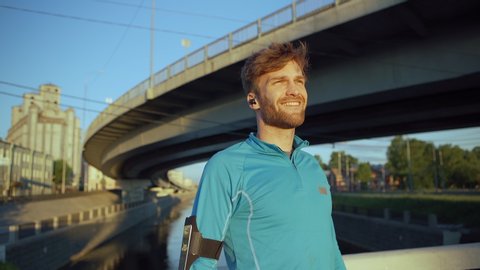 Lockdown medium shot of handsome young man finishing jogging cardio workout, looking away, catching breath and smiling standing on river over bridge. Headphones in his ears, cellphone armband on arm