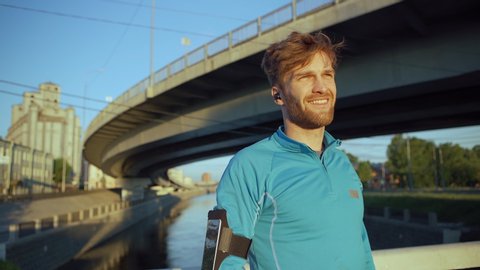 Handsome young athlete leaning on bridge railing while catching breath and recovering after jogging workout. Smiling man looking away and thinking in evening light