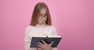 Thoughtful girl wearing eyeglasses and casual shirt reading new book and underlining interesting phrases with pen. Isolated over pink studio background.