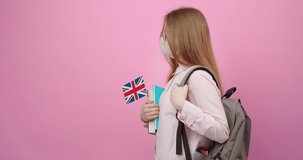 Pretty young girl in eyeglasses and medical mask with backpack holding books and British flag. Isolated over pink studio background. Concept of education and foreign languages.