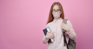 Portrait of female student with blond hair holding books in hand and backpack on shoulder over pink background. Smart woman wearing eyeglasses, casual shirt and protective medical mask.