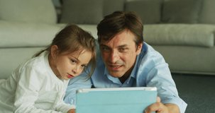 A happy smiling father and daughter enjoying time together and using a tablet for family entertainment while lying on a floor in living room at home.