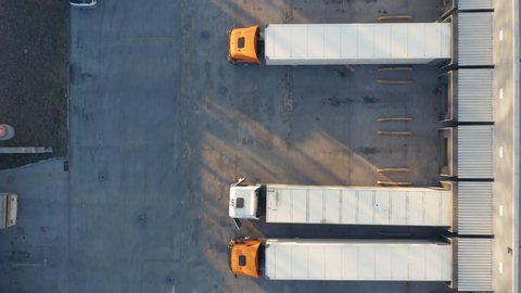 Aerial top down view of a lot of semi trucks with cargo trailers awaiting for loading/unloading goods on ramps on the big logistics park (loading hub) with warehouse.