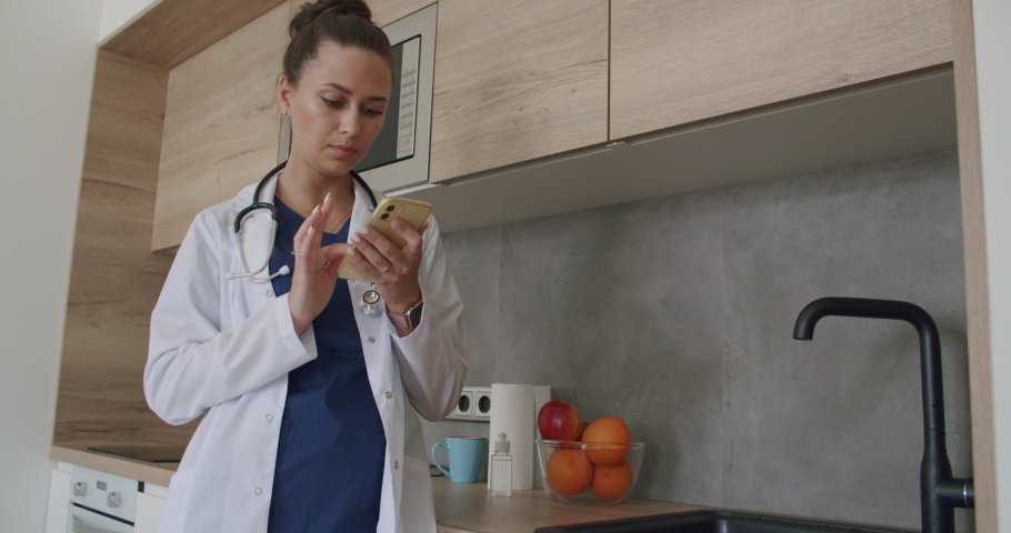 Lunch break in a medical facility. Female doctor in lab coat standing in clinic and scrolling on smartphone screen while kitchen in background. 4k raw video footage slow motion