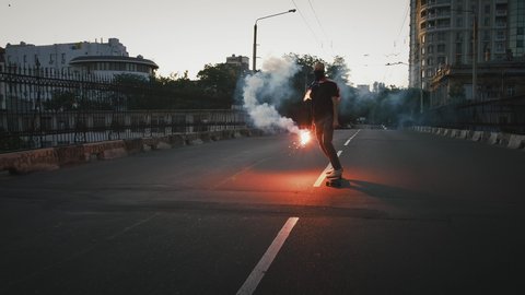 Young male in bandana on face and flag of USA tied on chest is skateboarding along deserted street with glowing red signal flare in hand. Slow motion
