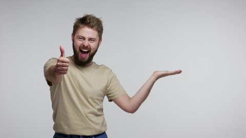 Surprised bearded guy pointing aside, holding empty space on palm and looking with open mouth amazed shocked, showing blank place for advertised product. studio shot isolated on gray background