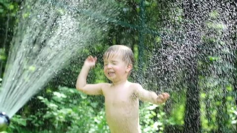 Cute toddler blond boy playing with garden sprinkler on summer hot day. Kid having fun on backyard jumping under spraying water from hosepipe. Happy childhood. Slow motion.