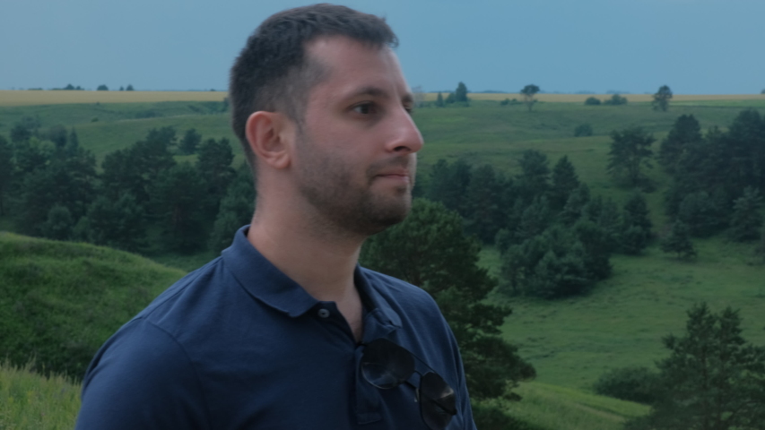 Close Up View Of Young Walking On A Hill And Looking Into The Distance. The Man Wearing Blue Polo. There Are Green Hills Around Him. Pensive Mood