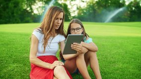 Cute girl and woman waving hands at camera on tablet computer outdoors. Positive mother and daughter using digital tablet for video call in park. Portrait of happy family chatting online in field