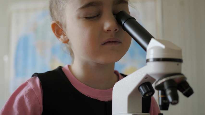 Little girl in science lab study samples under microscope. Schoolgirl looking through microscope in science class. Child looking into microscope, studing biology, chemistry in school laboratory.