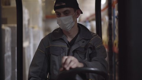 Portrait of confident serious Caucasian driver driving warehouse loader. Young handsome male employee in face mask working at factory storage on Covid-19 pandemic. Transporting, logistics.