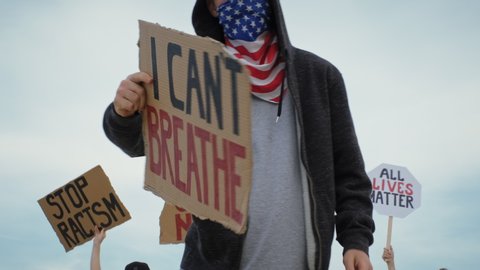 Young man on protest stands with sign All Lives Matter, on cardboard in hands, protest rally Black Lives Matter with group of people against blue sky. Riots in world. Protests in America, Europe