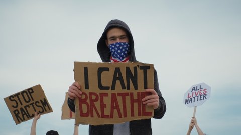 Young man on protest stands with signI Can't Breathe, on cardboard in hands, protest rally Black Lives Matter with group of people against blue sky. Riots in world. Protests in America, Europe