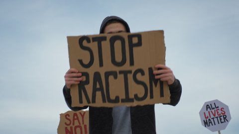 Young man on protest stands with sign Stop Racism, on cardboard in hands, protest rally Black Lives Matter with group of people against blue sky. Riots in world. Protests in America, Europe