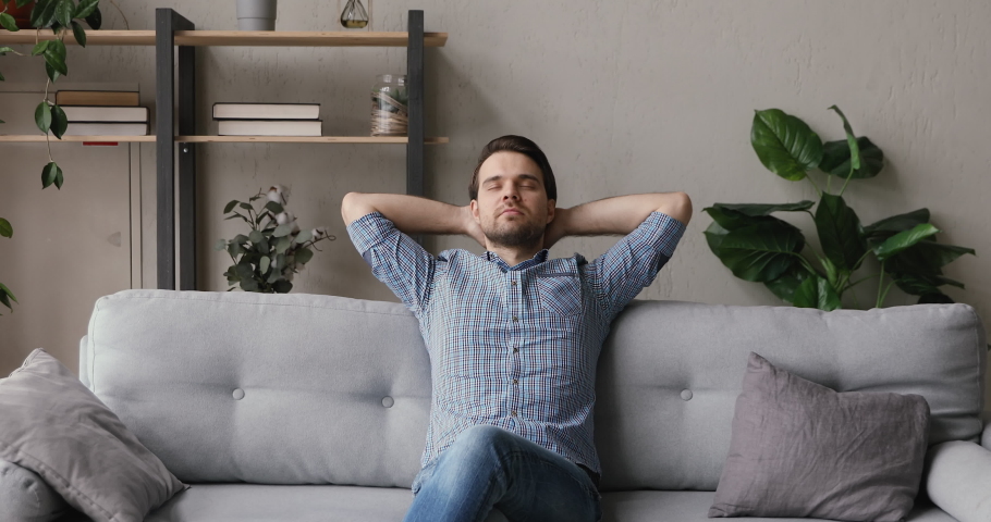 Serene man resting on couch puts hands behind head closed eyes enjoy fresh humidifier conditioned air. Male look into distance feels relaxed. New rented flat, homeowner, modern apartment owner concept Royalty-Free Stock Footage #1055363138