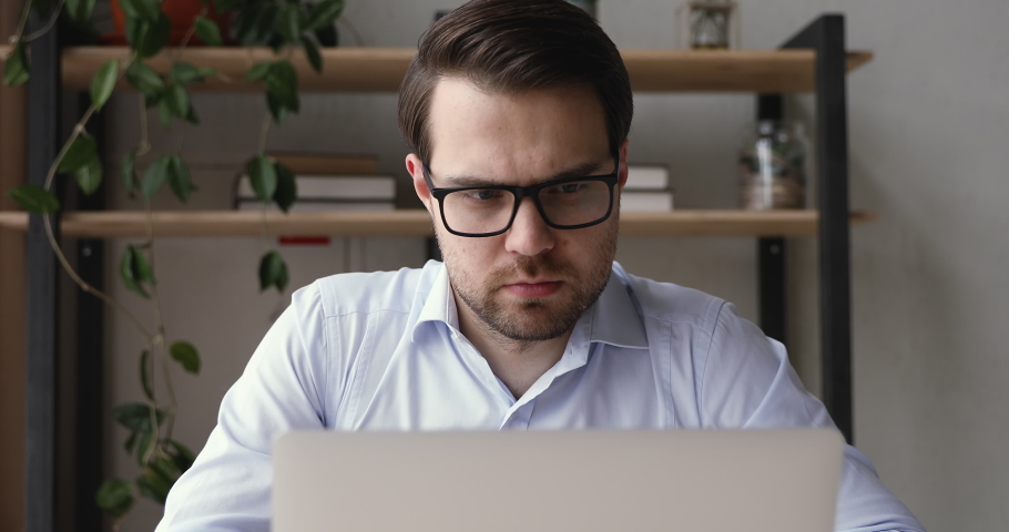 Millennial caucasian businessman sit at desk indoor wearing eyeglasses working using laptop learn new application having questions thinking search solution having fruitful busy workday in office room Royalty-Free Stock Footage #1055363162