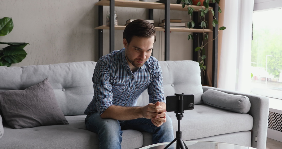 Man record videovlog using smart phone seated on couch alone in living room, put device on tripod guy film new vlog make on-line live stream, lead webinar distant chat, modern tech video event concept Royalty-Free Stock Footage #1055363315