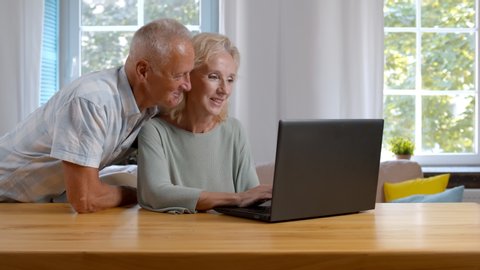 Portrait of mature woman sitting ant table and her husband hugging her from behind browsing internet together purchasing vacation tour or shopping online