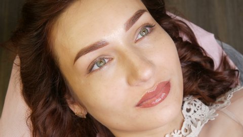 Beautiful young woman with permanent lip make-up and microblading eyebrow tattoo lies with a beauty parlor after the procedure. Close-up girl face.