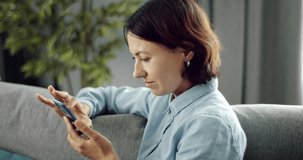 Side view of mature woman with dark hair spending leisure time at home and using smartphone. Charming lady in domestic outfit sitting on couch with modern gadget.