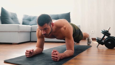 Young man exercising at home. Video of strong powerful guy stand in plank position on yoga mat. Confident concentrated man training in living room.