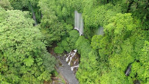 Aerial orbital shot around a high waterfall in a lush, green jungle, water falling forming a curtain than a watercourse on rocks continue as a river.